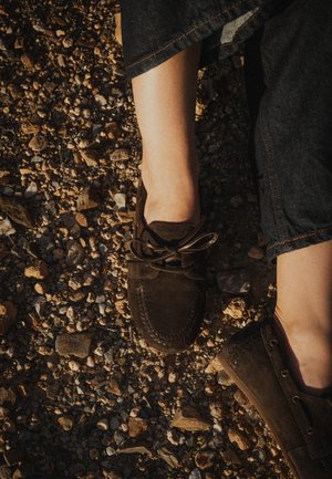 Pies con mocasines marrón oscuro sobre un suelo rocoso, persona vestida con pantalones de mezclilla azul oscuro.