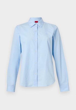 Cămașă albastru deschis cu mâneci lungi, cu nasturi și guler ascuțit, cu un mic logo pe pieptul stâng, afișată pe un fundal alb simplu.
