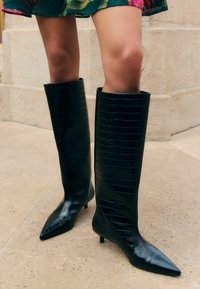 Knee-high melnas ādas zābaki ar spīdīgu krokodila rakstu, izteiksmīgiem pirkstiem un mazu papēdi, paredzēti pieguļoša izskata radīšanai.