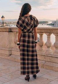 Abito lungo in tessuto scozzese con maniche corte a sbuffo, dettaglio a chiave sulla schiena e vita aderente. Indossato con stivaletti alla caviglia e braccialetti.