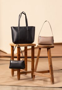 Tre håndtasker udstillet: en stor sort tote med en glat overflade, en lille sort clutch og en medium taupe skuldertaske, alle med enkle designs.