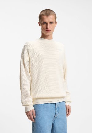 Joven con cabello corto que lleva un suéter de punto color crema y suelto, junto con jeans azul claro, de pie contra un fondo blanco liso.