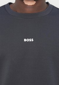 Gros plan d'une personne portant un t-shirt gris foncé avec le mot "BOSS" imprimé en blanc sur la poitrine.