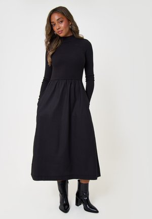 Femme portant une longue robe noire à manches longues et col montant, associée à des bottines noires brillantes, debout devant un fond blanc.