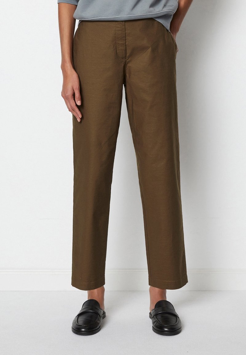 Personne portant un pantalon marron droit et des mocassins noirs sans lacets, debout contre un mur blanc avec les mains dans les poches.