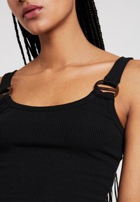 Débardeur noir à côtes avec bretelles réglables, mettant en valeur des éléments en métal. La vue rapprochée montre la texture et le détail des bretelles.