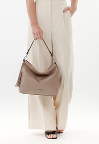 Sac à main en cuir tan avec un design décontracté, motif matelassé, quincaillerie en argent, pompon amovible et une bandoulière noire.