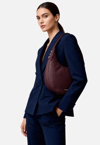 Borsa a tracolla in pelle bordeaux con forma curva, cuciture a contrasto e accessori in argento, indossata sopra un completo blazer blu navy.