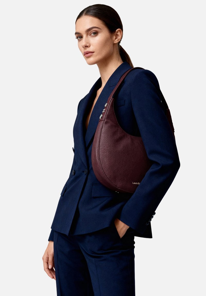 Borsa a tracolla in pelle bordeaux con forma curva, cuciture a contrasto e accessori in argento, indossata sopra un completo blazer blu navy.