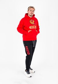 Felpa rossa con un grande logo dorato "49ERS" e un grafico del casco. Abbinata a pantaloni della tuta neri con stampa "49ERS", completata da sneakers bianche.