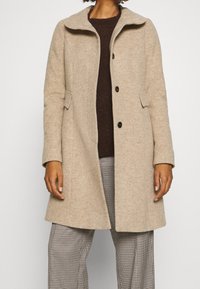 Abrigo de lana beige con cuello, cierre de botones, dos bolsillos laterales y mangas largas, combinado con un suéter marrón oscuro y pantalones de cuadros.