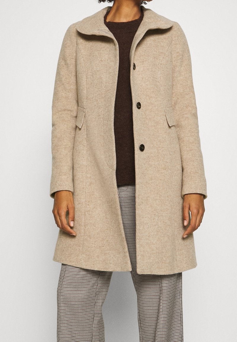 Abrigo de lana beige con cuello, cierre de botones, dos bolsillos laterales y mangas largas, combinado con un suéter marrón oscuro y pantalones de cuadros.