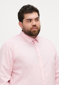 Camisa de botones de color rosa claro, hecha de una tela suave, con un cuello clásico y un corte relajado. Diseño simple sin patrones visibles.