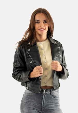 Chaqueta de motociclista negra de cuero corta con herrajes plateados, con cuello de muesca y bolsillos con cremallera. Llevada sobre una blusa beige de tejido peludo.