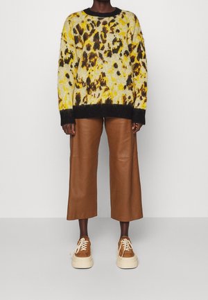 Personne portant un pull jaune et marron à taches, un pantalon en cuir marron large et court, et des baskets compensées marron avec des lacets crème.