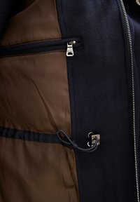 Laivaston takki, jossa on vetoketjullinen tasku, ruskea vuori ja kiristettävä yksityiskohta. Valmistettu pehmeästä materiaalista, jossa on sileä pinta.