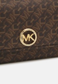 Ruda raštuota rankinė, pagaminta iš tekstūrinio medžiagos, su auksiniu apvaliu logotipu, kuriame yra "MK" ir "MICHAEL KORS" tekstas priekyje.