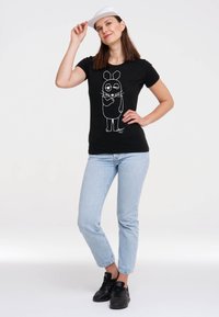 Black t-shirt featuring a white cartoon rabbit design, paired with light blue jeans and black sneakers. The model wears a white cap.