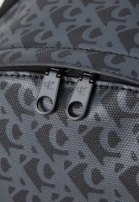 Šedý batoh se vzorem vyrobený z materiálu s charakteristickým logo. Černá zipová kovová s dvojicí tahátek, což zvyšuje jeho odolnost a funkčnost.