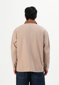 Man with black hair wearing beige jacket with a brown collar and dark blue jeans, standing facing away against a white background.