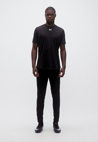 Ενήλικος άνδρας όρθιος, φοράει μαύρο αθλητικό μπλουζάκι, μαύρο παντελόνι και μαύρα ποδοσφαιρικά παπούτσια, μπροστά σε απλό λευκό φόντο.