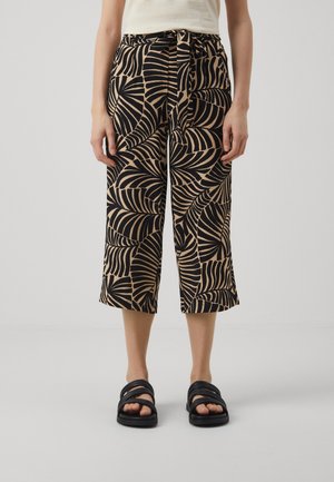 Vrouw draagt beige en zwart patroon cropped broek met een gestrikte taille en zwarte open teen sandalen, staand op een effen achtergrond.