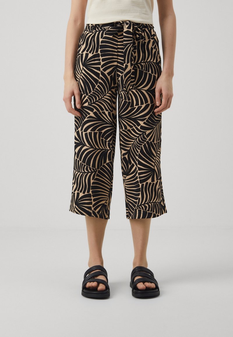Femme portant un pantalon court beige et noir à motifs avec une taille nouée et des sandales noires ouvertes, debout sur un fond uni.