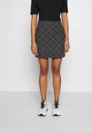 Vrouw draagt een zwart topje, grijze geruite A-lijn rok en witte sneakers, staand tegen een effen lichte achtergrond.