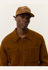 Brown textured jacket with two front pockets and button closure, paired with a tan cap featuring embroidered text and patterns.
