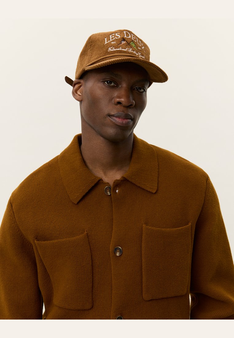 Brown textured jacket with two front pockets and button closure, paired with a tan cap featuring embroidered text and patterns.
