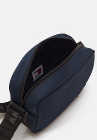 Mørkeblå stoff crossbody-veske med glidelås, som viser innvendig fôr og en logo-tag. Har en svart justerbar stropp.