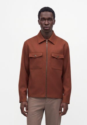 Ung mand iført en brun lynlåsjakke med to forlommer med klap og beige bukser mod en ensfarvet hvid baggrund.