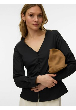 Kvinde med lyst hår iført en sort, langærmet bluse med knapper, holder en foldet brun ruskindsklædepung mod en ensfarvet baggrund.