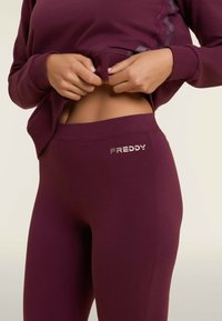 Set di abbigliamento attivo color bordeaux composto da una felpa aderente e leggings. Texture liscia con logo 'FREDDY' metallico sulla vita.