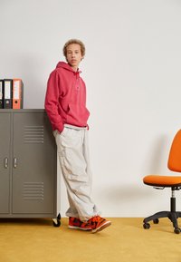 Ung person i röd hoodie och beige cargobyxor lutar sig mot en grå skåp med mappar bredvid en orange kontorsstol på en guldfärgad golv.