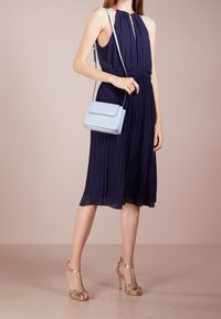 Marindrabbad plisserad klänning med halterneck, kombinerad med gyllene remsandaler och en ljusblå handväska. Slät tyg med ett flödande m önster.