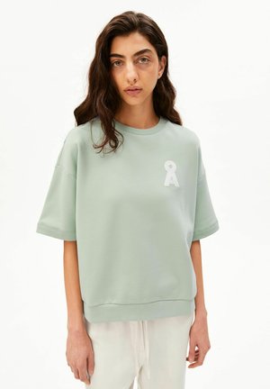 Femme aux longs cheveux foncés portant une chemise ample à manches courtes vert clair avec un logo blanc "OA", assortie à un pantalon de couleur claire.