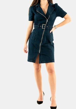 Femme portant une robe bleu foncé ajustée jusqu'aux genoux avec fermeture éclair à l'avant, ceinture, manches courtes et talons noirs, debout, une main sur la hanche.