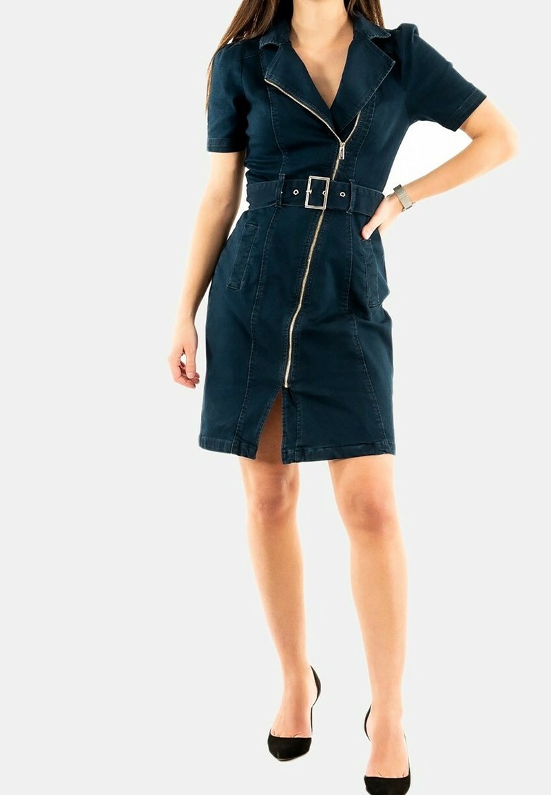 Femme portant une robe bleu foncé ajustée jusqu'aux genoux avec fermeture éclair à l'avant, ceinture, manches courtes et talons noirs, debout, une main sur la hanche.