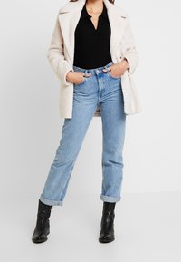 Beige, fluffig kappa över en svart, ribbad topp och ljusblå raka jeans, i kombination med svarta ankelstövlar, händerna i fickorna, neutral bakgrund.