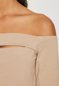 Beige off-shoulder topp med en tettsittende design, med et horisontalt utsnitt ved halsen og lange ermer. Myk, elastisk stoff.