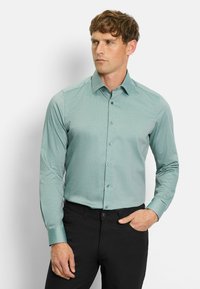 Hellgrünes Langarmhemd mit spitzem Kragen, knöpfbare Vorderseite und dezentem Texturmuster, kombiniert mit schwarzen Hosen.