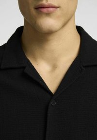 Czarna teksturowana koszula z krótkim rękawem, z wyciętym kołnierzem i pojedynczym czarnym guzikiem przy dekolcie, wykonana z miękkiej, lekko nieusystematyzowanej tkaniny.