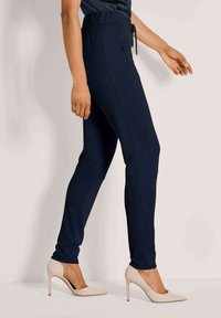 Vrouw in navy slim-fit broek met een zijrits zak en beige hoge hakken, staand tegen een lichte achtergrond.