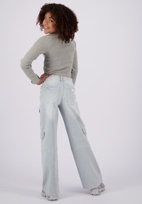 Maglia a maniche lunghe grigio chiaro abbinata a pantaloni larghi a righe di colore blu chiaro, con tasche cargo e una vestibilità rilassata. Modella di spalle che sorride.