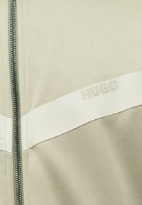 Ανοιχτό μπεζ ύφασμα με διαγώνια κρεμ λωρίδα που φέρει αχνό κείμενο "HUGO" και κάθετο μεταλλικό φερμουάρ στην αριστερή πλευρά.
