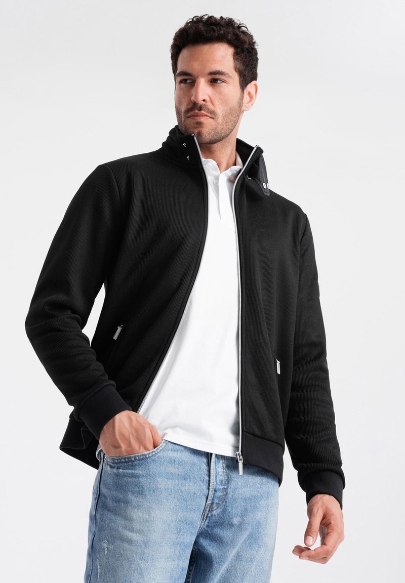 Giacca nera con zip, texture a coste, colletto alto e tasche laterali. Indossata sopra una camicia bianca, abbinata a jeans azzurri.
