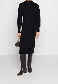 Robe tricot noire à manches longues avec col montant, taille ceinturée et poignets côtelés, associée à des bottes à talons hautes à genoux et un petit sac à main.