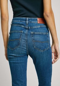Smala denimjeans i medelblå tvätt, med sydda bakfickor, läderlogotyp vid midjan och en mjuk yta.