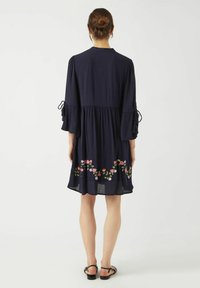 Granatowa sukienka z rękawami w kształcie dzwonu do łokci, zdobiona kolorowym haftem kwiatowym wzdłuż dołu oraz regulowanymi wiązaniami na rękawach.